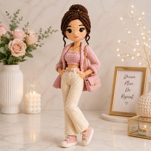 Può includere: Una bambola lavorata all'uncinetto a mano con capelli castani raccolti in uno chignon, che indossa un blazer rosa, un top corto rosa abbinato, pantaloni color crema con cintura e scarpe da ginnastica rosa. La bambola porta una borsa rosa.