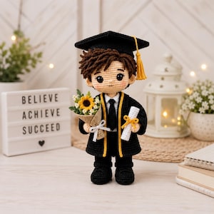 Può includere: Una bambola all'uncinetto vestita con una toga di laurea nera con bordi gialli, un tocco nero con nappa gialla, una camicia bianca e una cravatta nera. La bambola tiene in mano un piccolo bouquet di girasoli e un diploma bianco.