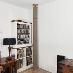 Può includere: Un tiragraffi cilindrico e alto per gatti, rivestito in un materiale strutturato marrone chiaro, si estende dal pavimento al soffitto. È posizionato accanto a una libreria bianca piena di CD e dischi in vinile. Un tavolo di legno con una lampada e una sedia sono in primo piano.