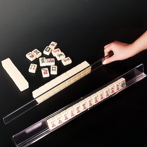 May include: A set of ivory-colored Mahjong tiles with red and black markings, displayed with clear acrylic tile racks. Some tiles are scattered on a black surface, with a hand reaching for a rack. The game set is designed for strategic gameplay.