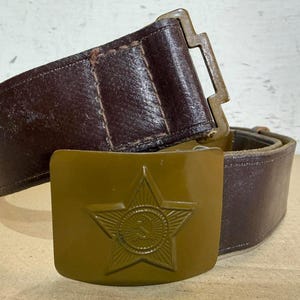 May include: A dark brown leather belt featuring a rectangular olive green metal buckle. The buckle is embossed with a prominent five-pointed star containing a hammer and sickle emblem in the center. The belt includes a metal loop and stitching.