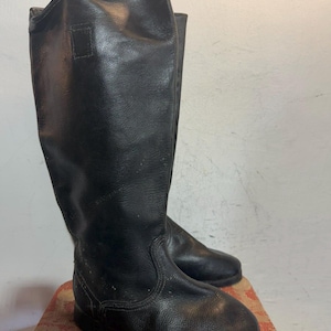 Puede incluir: Un par de botas negras hasta la rodilla con acabado texturizado, punta redondeada, tacón bajo y costuras visibles a lo largo del pie y la caña. Las botas presentan una silueta alta y estructurada que llega hasta la zona de la pantorrilla.