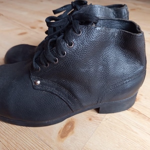 Puede incluir: Un par de botines negros con parte superior de cuero texturizado, punta redondeada y cierre de cordones negros. Los botines tienen un tacón bajo y un detalle de remache de metal plateado visible cerca de la base del sistema de cordones.