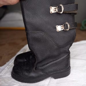 May include: A pair of black leather boots with two adjustable straps and silver-colored buckles. The boots have a textured surface and a sturdy sole. The boots are knee-high.