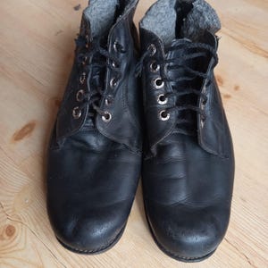 Puede incluir: Un par de botines de cuero negro con punta redondeada, ojales metálicos plateados y cordones negros. El interior de las botas está forrado con un material polar gris y texturizado, lo que proporciona un contraste visible con el exterior negro liso.