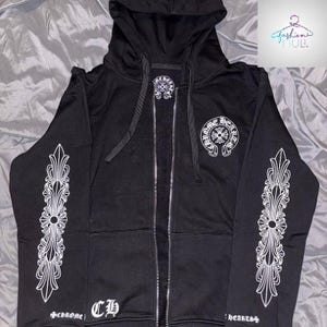 May include: Black zip-up hoodie with white decorative designs on the sleeves and a logo on the chest. The hoodie features a drawstring hood and a full-length zipper. The text "Chrome Hearts" is visible on the front.