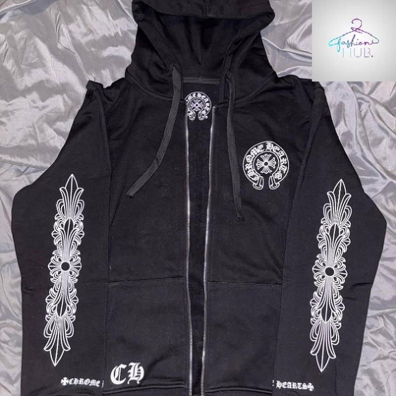 May include: Black zip-up hoodie with white decorative designs on the sleeves and a logo on the chest. The hoodie features a drawstring hood and a full-length zipper. The text "Chrome Hearts" is visible on the front.