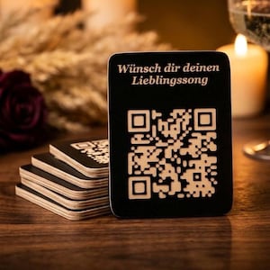 Könnte beinhalten: Ein Stapel schwarzer Untersetzer mit hellem Rand und QR-Code-Design. Ein Untersetzer steht aufrecht und zeigt den Text "Wünsch dir deinen Lieblingssong" in heller Farbe. Die Untersetzer liegen auf einer Holzoberfläche.