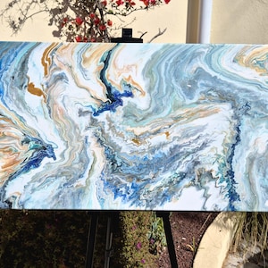 May include: Abstract acrylic painting on canvas. The artwork features a fluid design with swirling patterns in shades of blue, white, gold, and teal. The painting is displayed on an easel outdoors, showcasing the vibrant colors and textures.
