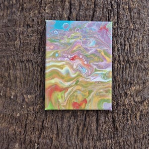 May include: A small, rectangular abstract painting on canvas. The artwork features a fluid, colorful design with shades of blue, purple, yellow, green, and red. The paint appears to have been poured and swirled, creating a unique, textured effect.