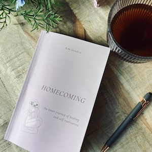 Könnte beinhalten: Ein Buch mit dem Titel "HOMECOMING" von B.M. Hendrix, mit dem Untertitel "Eine innere Reise der Heilung und Selbstverwirklichung". Ein Stift und ein Glas Tee liegen neben dem Buch. Das Buchcover ist helllila mit einer Strichzeichnung einer Person.