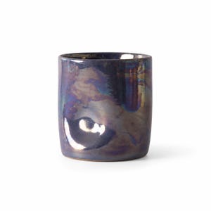 May include: A cylindrical ceramic cup featuring a deep, iridescent glaze with swirling metallic tones of dark purple, blue, and bronze. The surface has a smooth, reflective finish with subtle indentations along the side and a wide, open rim.