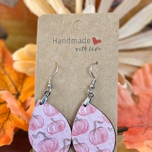 Puede incluir: Pendientes en forma de lágrima con un diseño de calabazas rosas. Se exhiben en una tarjeta marrón con la frase "Handmade with love". El fondo incluye hojas otoñales en tonos naranja y amarillo.