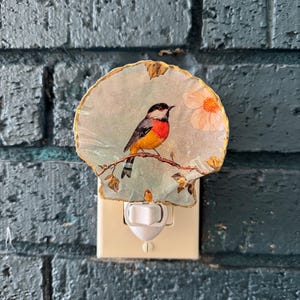 May include: A decorative night light featuring a painted bird perched on a branch with a flower. The bird has a black head, red chest, and yellow belly. The night light is plugged into a wall outlet against a blue brick wall.