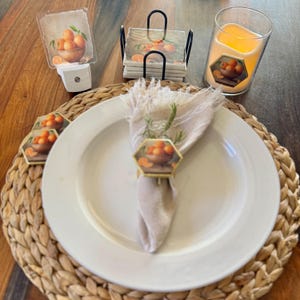 May include: A table setting featuring a white plate, napkin, and napkin ring. The napkin ring and other items have a design of oranges in a wooden bowl. Other items include a candle, coasters, and a night light.