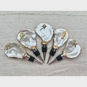 Pode incluir: Cinco rolhas de garrafa decorativas com tampas brancas peroladas e detalhes dourados. Cada rolha apresenta um charme dourado diferente: uma casa, uma estrela, uma figura, um pássaro e um desenho floral. As rolhas têm vedações de borracha preta e bases de metal prateado.