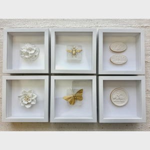 May include: Six white framed shadow boxes with decorative elements. The boxes contain a white flower, a gold bee, two white oval reliefs, a white flower, a gold butterfly, and a white round relief.