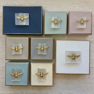 Könnte beinhalten: Ein Set aus drei Keramikbechern mit gesprenkelter, matter Oberfläche. Die Becher sind in Creme-, Hellgrau- und Dunkelgrautönen gehalten. Jeder Becher hat ein schlichtes, minimalistisches Design und einen abgerundeten Henkel.