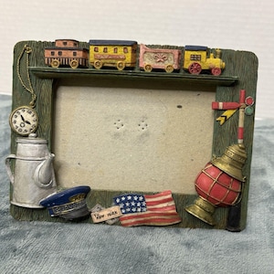 Op de afbeelding: Decoratieve fotolijst met een treinthema. De lijst heeft miniatuurwagons, een pet van een conducteur, een Amerikaanse vlag en een rode lantaarn. De lijst is gemaakt van hout en heeft een groen-bruin kleurenschema.