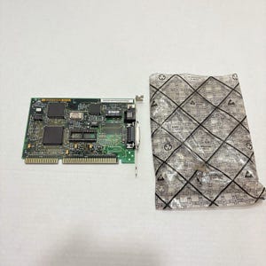 May include: A green printed circuit board network interface card with various black integrated circuits, gold-plated edge connectors, and a metal bracket, positioned next to a translucent anti-static plastic bag with a black grid pattern.