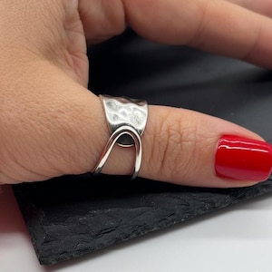 May include: A silver ring with a hammered texture and a distinctive curved design. The ring is worn on a finger with red nail polish. The ring's design is modern and eye-catching.