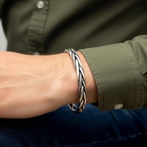 May include: Silver-toned braided cuff bracelet. The bracelet features a woven design with a textured appearance. The jewelry is worn on a wrist, against a background of a green shirt and blue jeans.