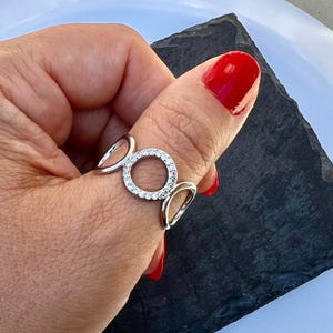 Peut inclure: Une bague argentée avec un motif circulaire incrusté de petites pierres précieuses transparentes. La bague comporte deux accents en forme de goutte de chaque côté du cercle de pierres précieuses. La bague est portée sur un doigt avec du vernis à ongles rouge.