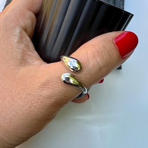 Könnte beinhalten: Ein silberner Ring mit zwei tropfenförmigen Elementen. Der Ring umschließt den Finger, wobei sich die Tropfen auf beiden Seiten befinden. Die Tropfen haben eine polierte, reflektierende Oberfläche. Der Finger trägt roten Nagellack.