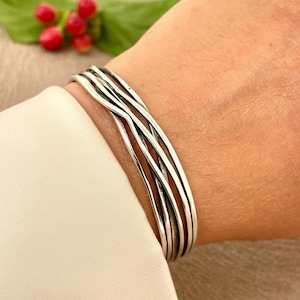 May include: A silver-toned bracelet with a woven design is shown on a wrist. The bracelet is made of multiple strands of metal, creating a textured appearance. Red berries and green leaves are visible in the background.