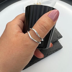 Peut inclure: Une bague en argent avec un design à double bande. La bague a une finition lisse et polie et une forme ondulée. La bague est portée sur un pouce. Le fond est blanc avec un objet noir.