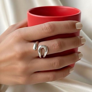 May include: A silver ring featuring a horseshoe design. The ring is worn on a finger, with the hand holding a red mug. The ring has a smooth, polished finish and a contemporary style.