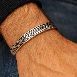 May include: A silver cuff bracelet with a detailed leaf pattern. The bracelet is worn on a wrist, showcasing the intricate design. The metal has a polished finish, reflecting light. The bracelet is a piece of jewelry.