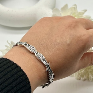May include: A silver bracelet featuring a repeating wing design. The bracelet is worn on a wrist, showcasing the intricate details of the feather-like patterns. The jewelry is made of a shiny, metallic material.