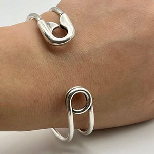 May include: A silver-toned cuff bracelet featuring a safety pin design at the top and a circular loop at the bottom. The bracelet is worn on a person's wrist, showcasing its unique, modern style. The metal has a polished finish.