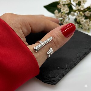 May include: A silver ring with a contemporary, geometric design. The ring has a straight bar and a curved, open band, creating a unique accessory. The ring is shown on a finger with red nail polish. The ring is a stylish piece of jewellery.