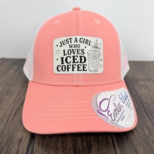 May include: A coral-colored trucker hat with a white mesh back and a rectangular patch on the front. The patch features black text that reads JUST A GIRL WHO LOVES ICED COFFEE and an illustration of a coffee cup with a bow. A sticker is on the brim.