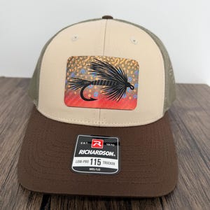 May include: A tan and brown trucker hat featuring a rectangular patch with a black fly fishing lure graphic over a colorful speckled background. The hat has a brown brim and a mesh back, with a black sticker on the brim reading Richardson 115.