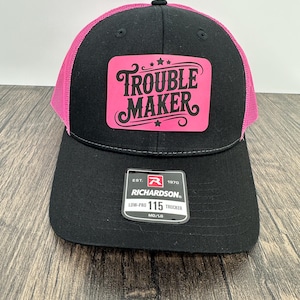 May include: Black trucker hat with a pink mesh back and a black brim. The front features a pink patch with the words "TROUBLE MAKER" in a stylized font. The hat has a Richardson tag.