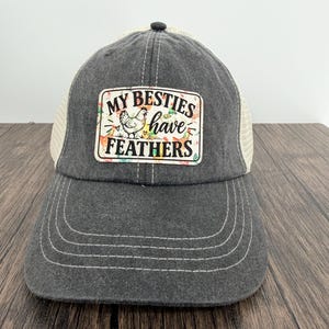 May include: A charcoal gray baseball cap with a mesh back and a rectangular patch on the front. The patch features a white background with a colorful chicken illustration and the black text "MY BESTIES have FEATHERS" with small heart accents.
