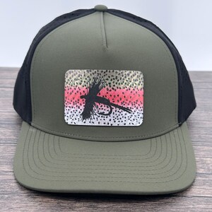 May include: A green and black baseball cap with a curved brim. The cap features a rectangular patch with a colorful trout pattern and a black fly fishing lure silhouette. The cap has a mesh back for ventilation.