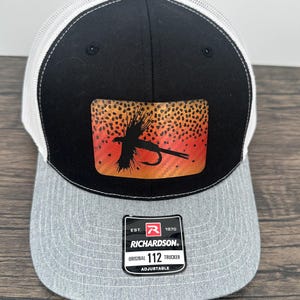 May include: A black, white, and gray trucker hat with a patch featuring a fly fishing lure design. The patch has a black silhouette of a fly and hook against a speckled orange and yellow background. The hat has a Richardson 112 tag.