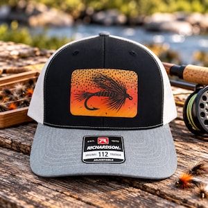 May include: A black and grey trucker hat featuring a rectangular patch with an orange and black speckled background and a black fly fishing lure graphic. The brim has a sticker that reads EST. 1970 RICHARDSON ORIGINAL 112 TRUCKER ADJUSTABLE.