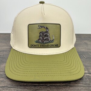 May include: A beige and olive green baseball cap featuring a patch with a coiled rattlesnake and the text "DON'T TREAD ON ME." The cap has a curved brim and a mesh back for ventilation. The patch is rectangular and has a green background.