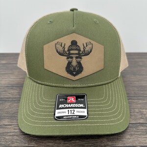 May include: Olive green and tan trucker hat with a patch featuring a moose wearing sunglasses and a knit cap. The hat has a curved brim and an adjustable closure. The Richardson brand label is visible.
