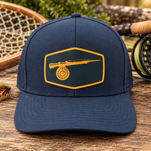 May include: A navy blue baseball cap featuring a hexagonal patch on the front. The patch is dark blue with a yellow border and displays a yellow graphic illustration of a fly fishing reel and rod. The cap has perforated side panels.