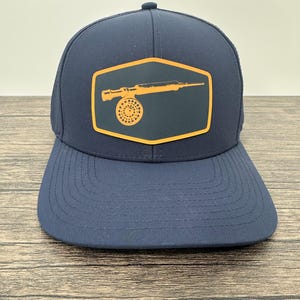 May include: Navy blue baseball cap with a structured crown and a curved brim. The cap features an orange patch with a fishing reel design. The patch is outlined in orange and is centered on the front of the cap. The cap is made of a durable material.