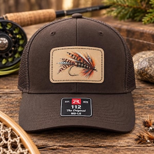 May include: A dark brown trucker hat featuring a tan rectangular patch with a detailed illustration of a colorful fishing fly. The brim displays a black sticker with the text Est. 1970 R 112 The Original MD-LG.