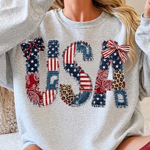 May include: A light gray sweatshirt with the word "USA" appliqued on the front. The letters are made of patchwork denim, paisley, leopard print, and star-spangled fabric, with red, white, and blue bows.