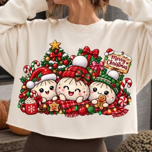 May include: A cream-colored sweatshirt featuring a festive graphic of three smiling dumpling characters wearing winter hats. The design includes a Christmas tree, wrapped gifts, a red mug, candy canes, and a sign that reads Merry Christmas.