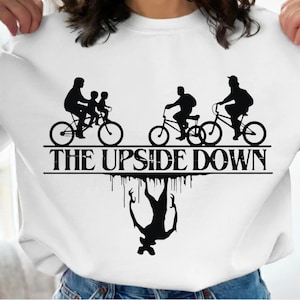 Puede incluir: Sudadera blanca con un gráfico negro de personas montando en bicicleta sobre el texto "THE UPSIDE DOWN". Debajo, una silueta de un monstruo. El diseño es una referencia a una serie de ciencia ficción popular.